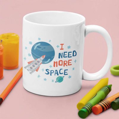 A photograph of a kids mug on a table surrounded by crayons ith the words I need more space printed on it alone with planets stars and moons