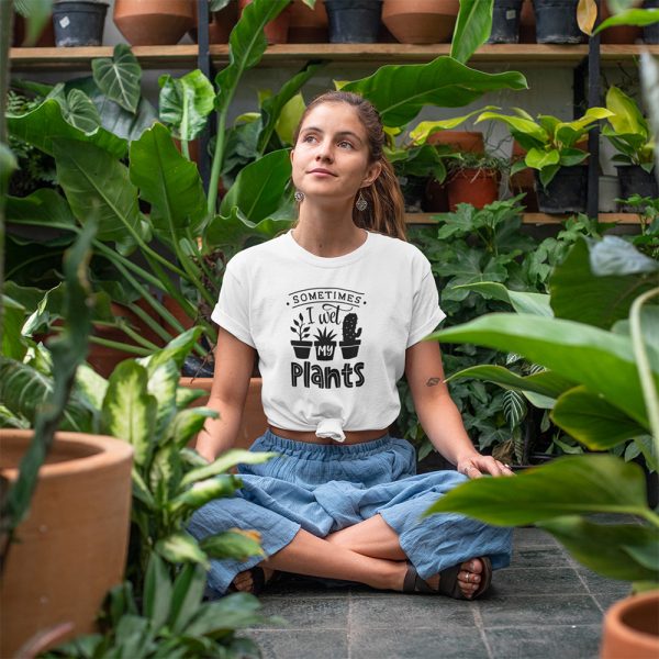 A Photo of a lady surrounded by plants in a yoga pose wearing a white t shirt with the words Sometimes I Wet My Plants printed on it along with images of potted plants