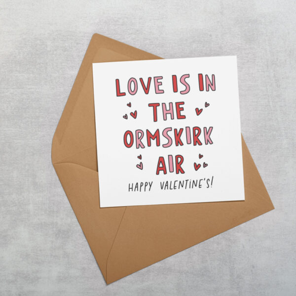 a white square greetings card with the wors love is in the ormskirk air happy valentines day, it is colourful with red and pink colours and love hearts and also the font is square shaped with sharp edges