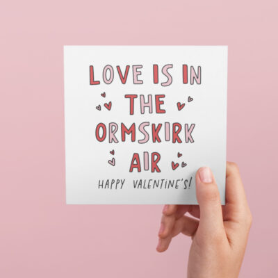 a white square greetings card with the wors love is in the ormskirk air happy valentines day, it is colourful with red and pink colours and love hearts and also the font is square shaped with sharp edges